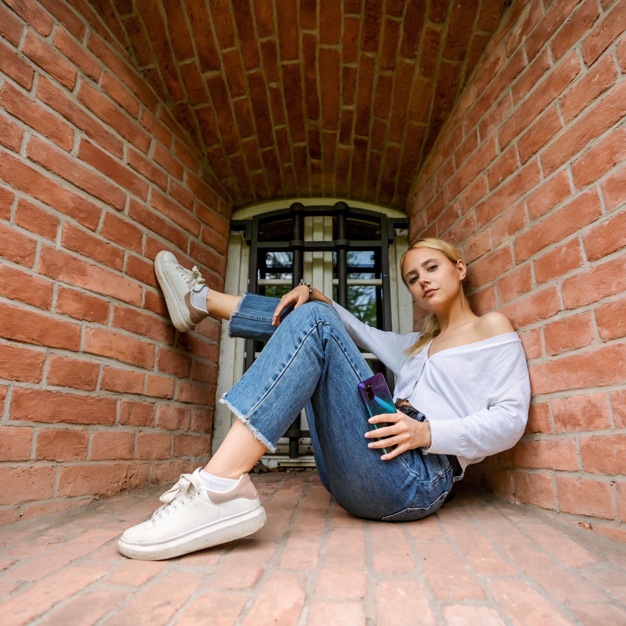 Młoda kobieta w białej bluzce i dżinsach siedzi w niszy z cegły, trzymając telefon komórkowy i patrząc w obiektyw.