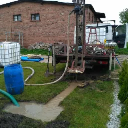 Wiercenie studni przy użyciu wiertnicy zamontowanej na samochodzie ciężarowym, w tle budynek z czerwonej cegły i białe zbiorniki na wodę.