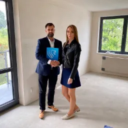 Mężczyzna w garniturze i kobieta w skórzanej kurtce uściskiem dłoni finalizują transakcję w surowym wnętrzu mieszkania z widokiem na zieleń.