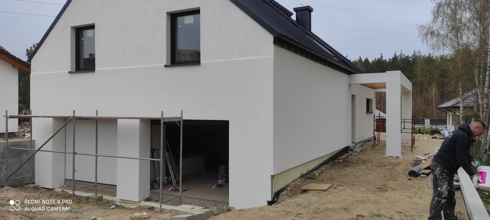 Nowoczesny dom jednorodzinny w Chojnicach po malowaniu elewacji na biało, widoczne rusztowania i pracownik przygotowujący się do dalszych prac wykończeniowych.