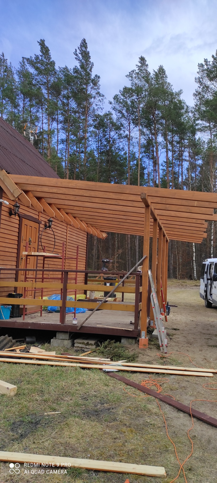 Drewniana konstrukcja zadaszenia tarasu w trakcie budowy przy domu z drewnianą elewacją, widoczne elementy rusztowania, drabina i narzędzia, tło stanowią drzewa.