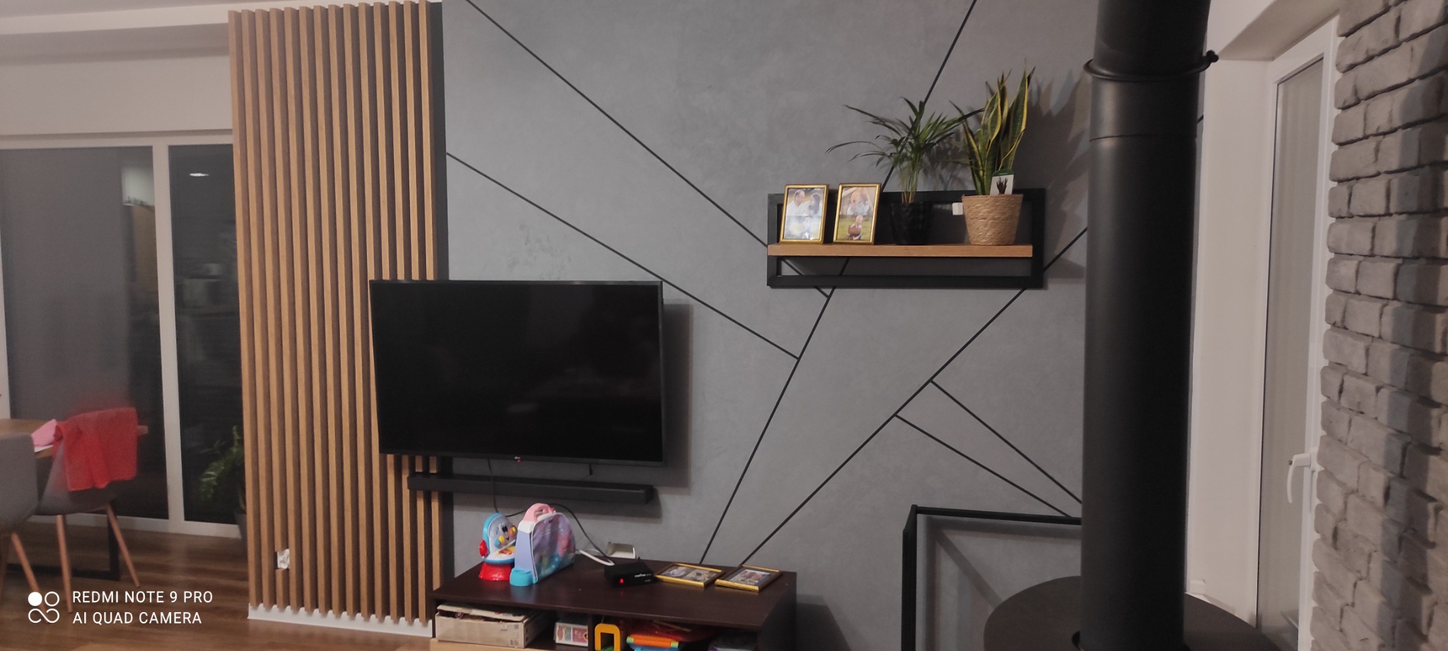 Szara ściana z geometrycznym wzorem, drewniane lamele dekoracyjne, czarny komin pieca wolnostojącego, półka z ramkami i roślinami doniczkowymi, telewizor na ścianie, ceglana ściana po prawej.