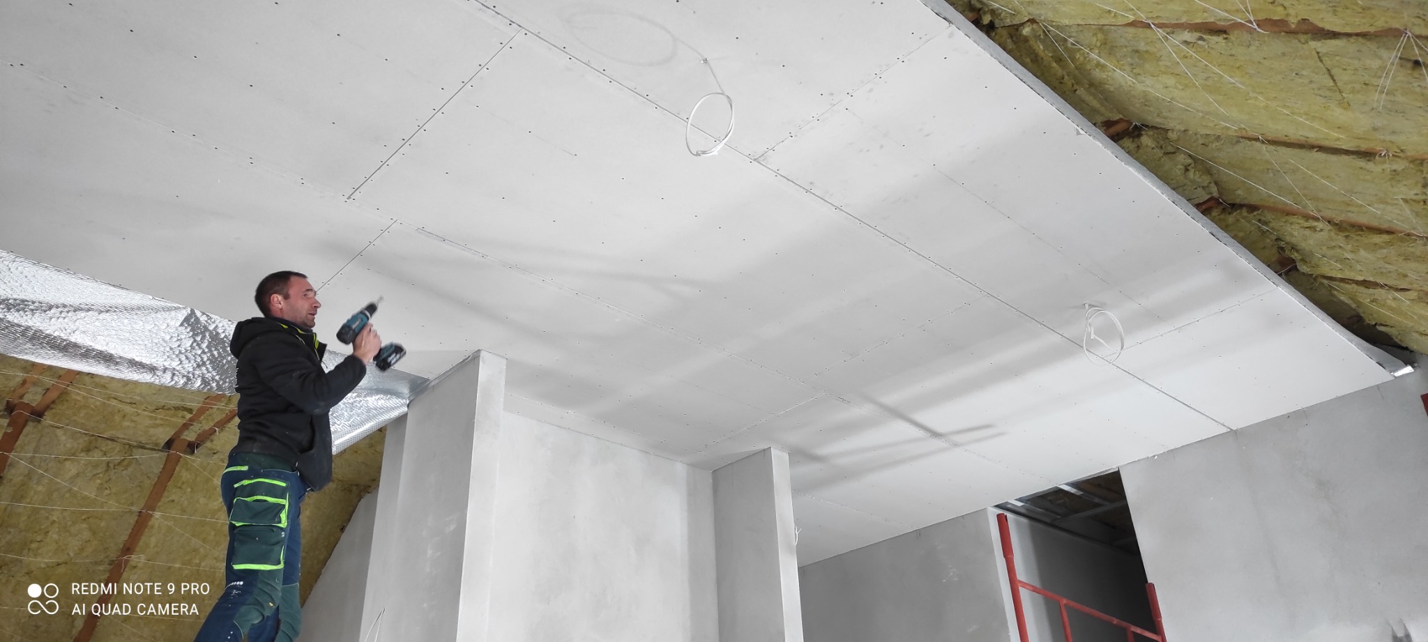 Pracownik montuje płytę kartonowo-gipsową na suficie poddasza, widoczne ocieplenie wełną mineralną i folią aluminiową, przewody elektryczne wystające z sufitu.