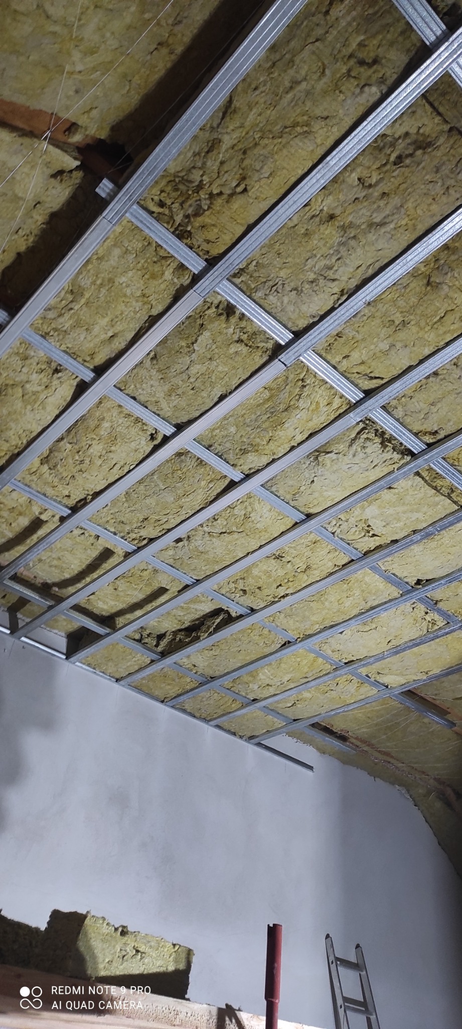 Sufit podwieszany z metalowym stelażem wypełnionym wełną mineralną, widok z dołu w trakcie montażu, fragment drabiny i czerwonego narzędzia w dolnej części kadru.