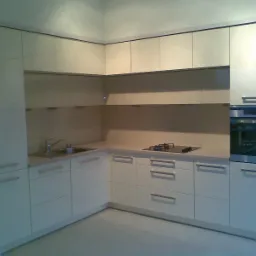 Jasna, narożna zabudowa kuchenna z lakierowanymi frontami, zintegrowanym piekarnikiem i płytą gazową, widok na fragment ściany w kolorze błękitu.