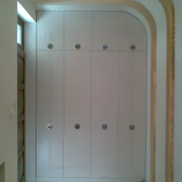 Zamknięta, biała szafa wnękowa z okrągłymi uchwytami, wkomponowana w łukowate przejście, oświetlona światłem punktowym.