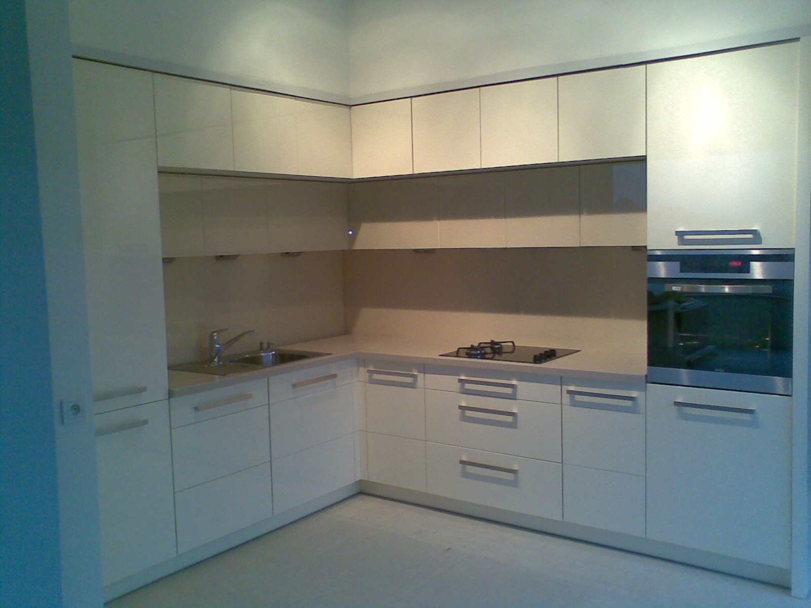 Jasna, narożna zabudowa kuchenna z lakierowanymi frontami, zintegrowanym piekarnikiem i płytą gazową, widok na fragment ściany w kolorze błękitu.