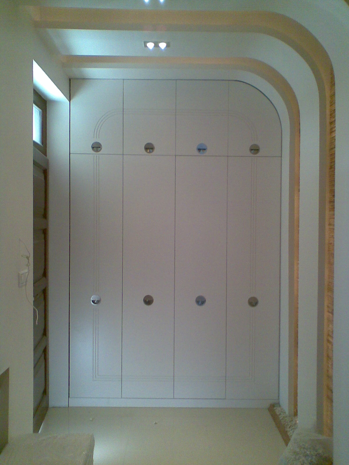 Zamknięta, biała szafa wnękowa z okrągłymi uchwytami, wkomponowana w łukowate przejście, oświetlona światłem punktowym.