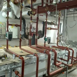 Rozbudowana instalacja hydrauliczna z rurami w kolorze ceglastym, manometry, zawory i elementy sterujące zamontowane na urządzeniu mechanicznym w pomieszczeniu technicznym.