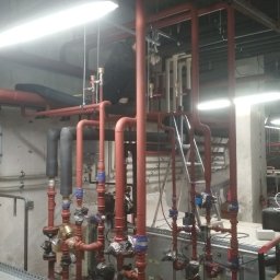 Vitali Paulouski - Rozbudowana instalacja grzewcza z licznymi rurami w kolorze czerwonym, zaworami z niebieskimi elementami i szarym zbiornikiem wyrównawczym na tle surowej ściany w piwnicy.