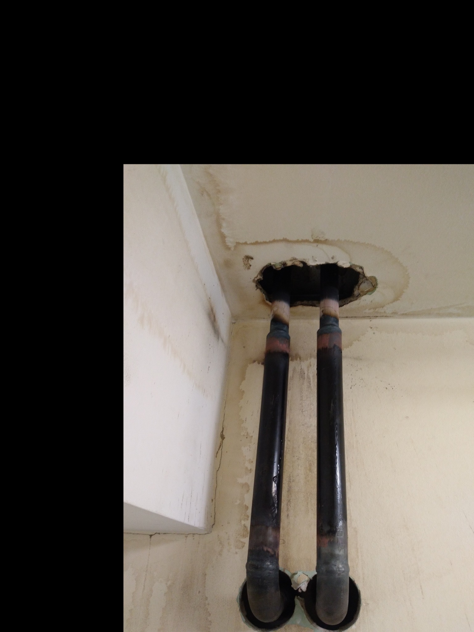 Dwie czarne rury wychodzące z dziury w suficie z widocznymi śladami wilgoci i zaciekami na ścianach.