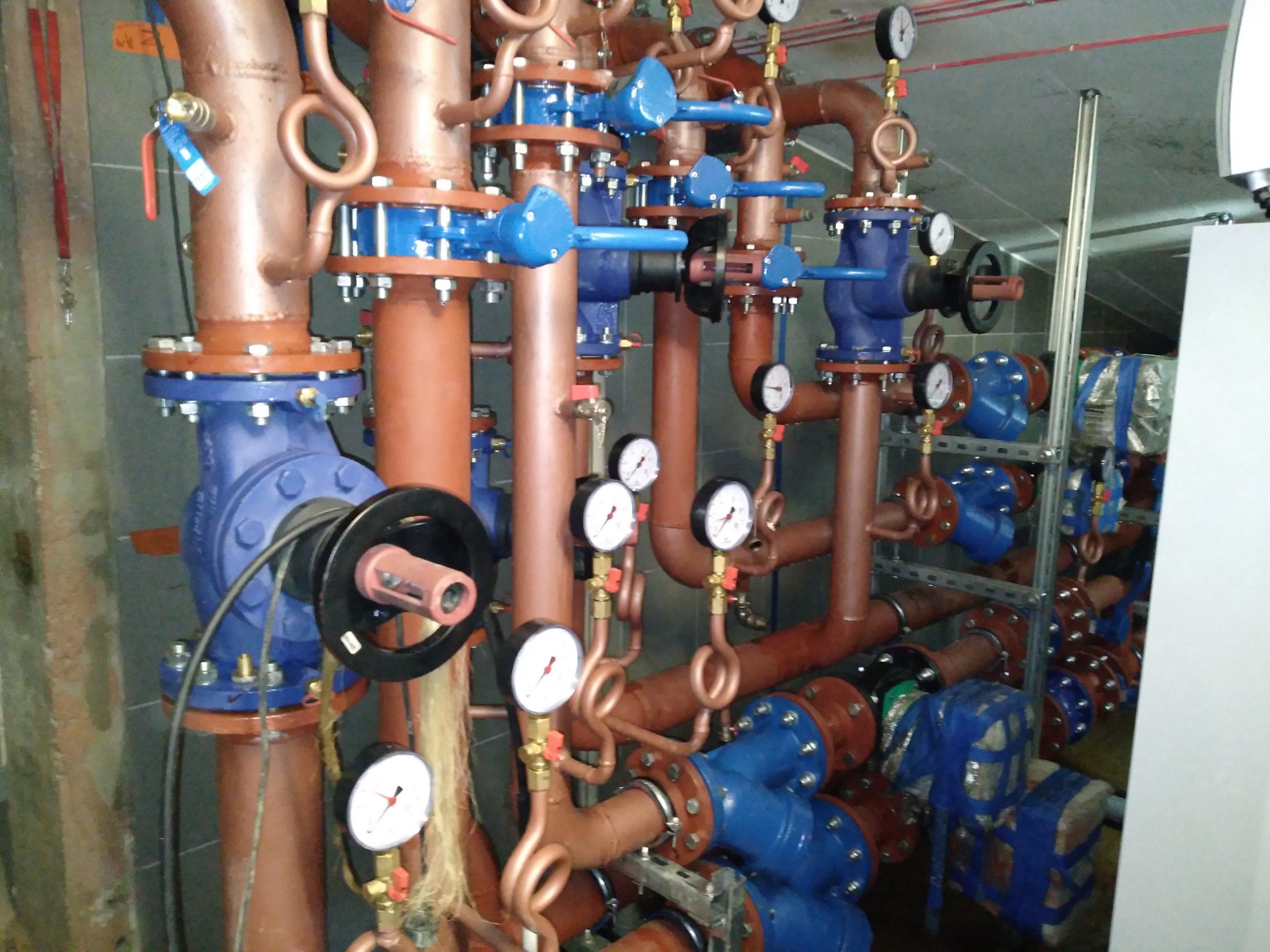 Złożona instalacja rurowa z zaworami, manometrami i elementami izolacyjnymi w pomieszczeniu technicznym.