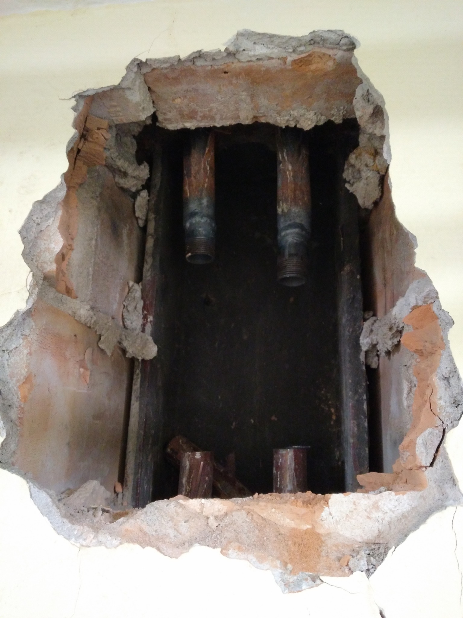 Otwór w ścianie odsłaniający rury instalacji wodnej z gwintowanymi końcówkami, otoczone gruzem i fragmentami cegieł.