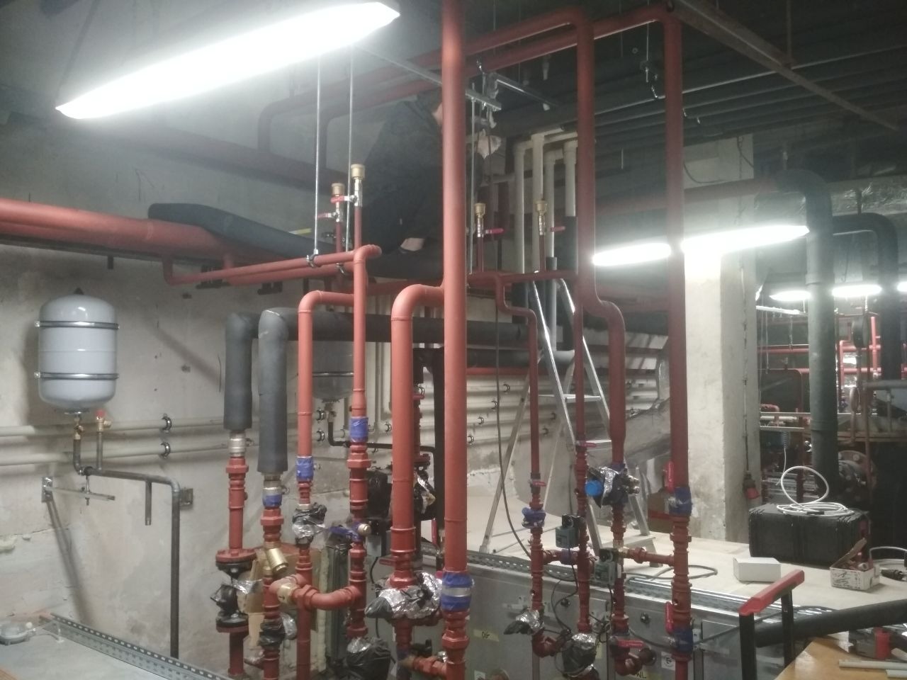 Rozbudowana instalacja grzewcza z licznymi rurami w kolorze czerwonym, zaworami z niebieskimi elementami i szarym zbiornikiem wyrównawczym na tle surowej ściany w piwnicy.