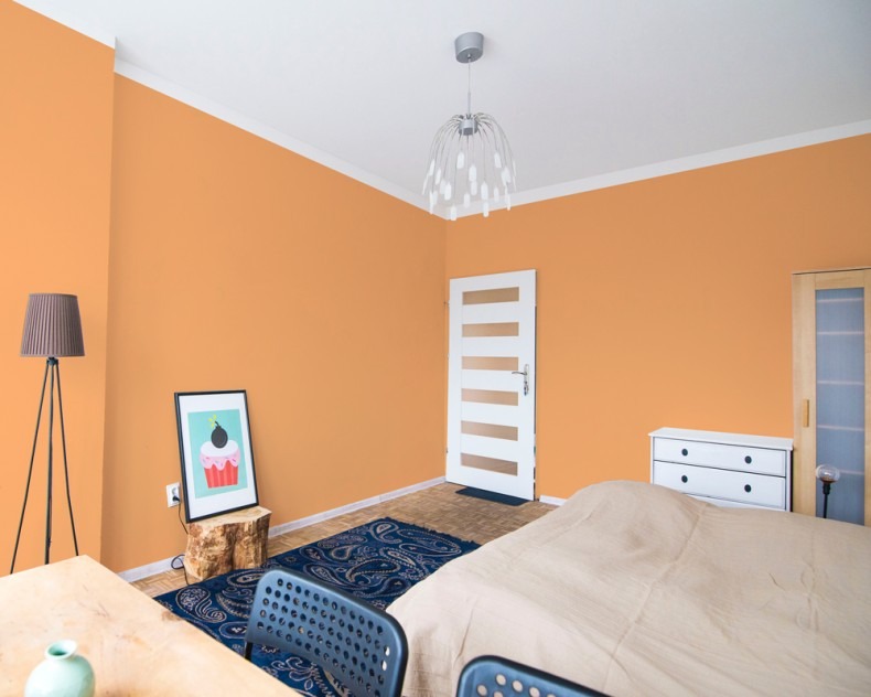 Pomarańczowe ściany w sypialni po malowaniu, widoczna biała stolarka drzwiowa z poziomymi przeszkleniami, lampa podłogowa na trójnogu, plakat z babeczką na pniu drzewa.