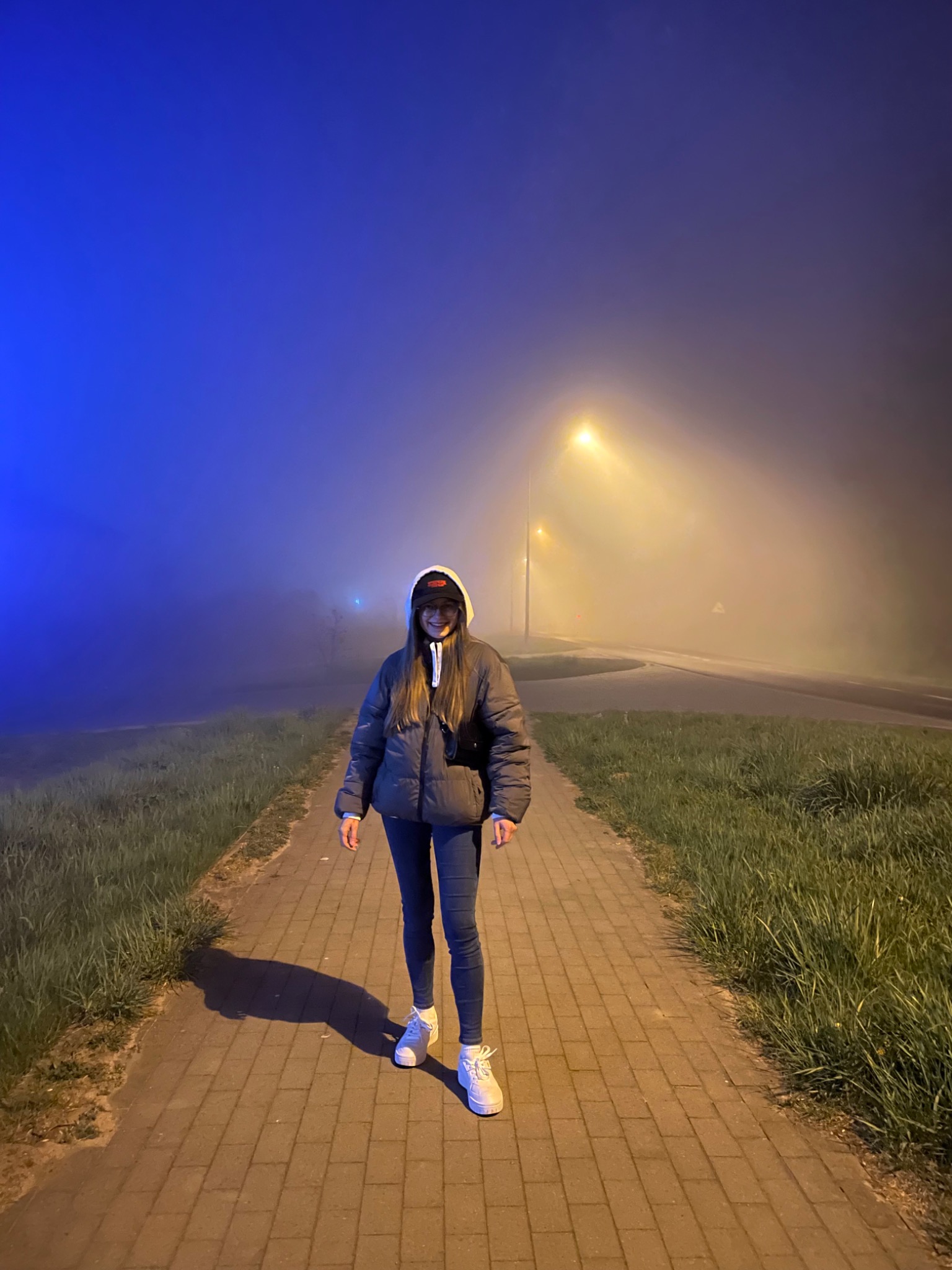 Kobieta w kurtce i czapce z daszkiem stoi na chodniku w gęstej mgle, oświetlona przez latarnie uliczne o ciepłej i chłodnej barwie.