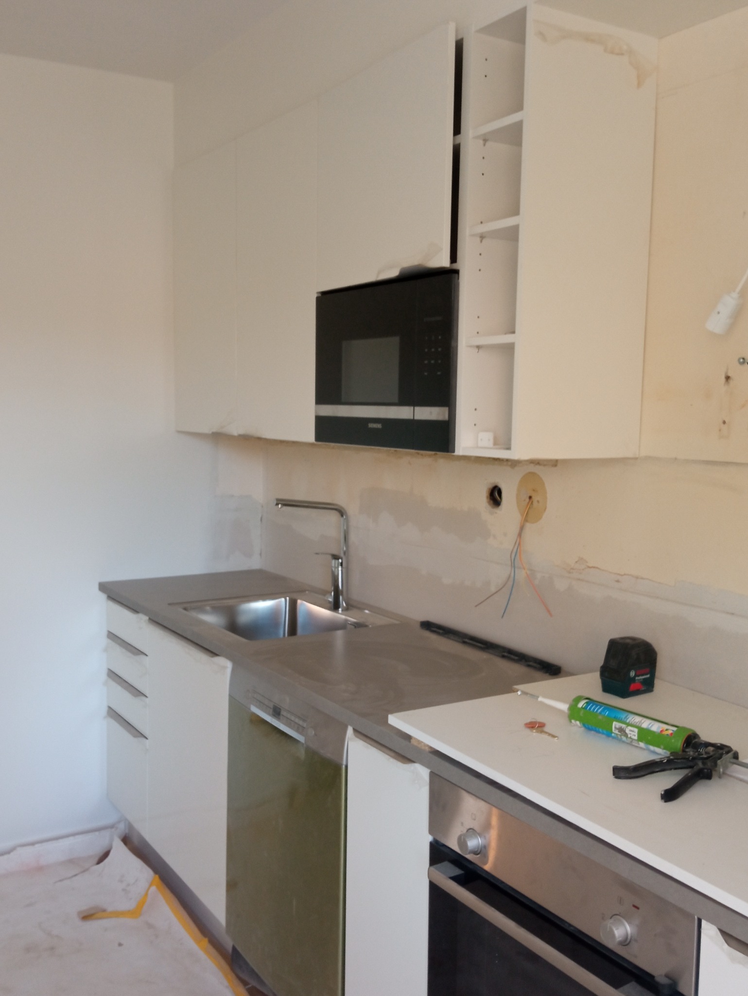 Wnętrze kuchni w trakcie montażu: białe szafki wiszące z wbudowaną mikrofalówką, szary blat ze zlewem, widoczne narzędzia i materiały montażowe, nieukończone ściany z wystającymi przewodami.