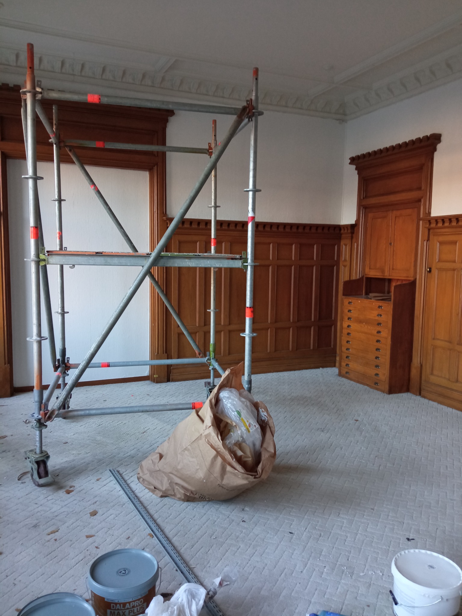 Wnętrze pokoju z drewnianą boazerią w trakcie remontu, widoczne rusztowanie, wiadro z farbą i worek z odpadami.
