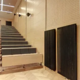 Aula z drewnianymi schodami i panelami akustycznymi na ścianach, dwa czarne grzejniki płytowe na ścianie, parkiet ułożony we wzór jodełki.