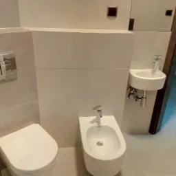 Nowoczesna łazienka z wiszącą toaletą, bidetem i umywalką, wykończona w jasnych, beżowych płytkach ceramicznych z minimalistycznymi akcentami.