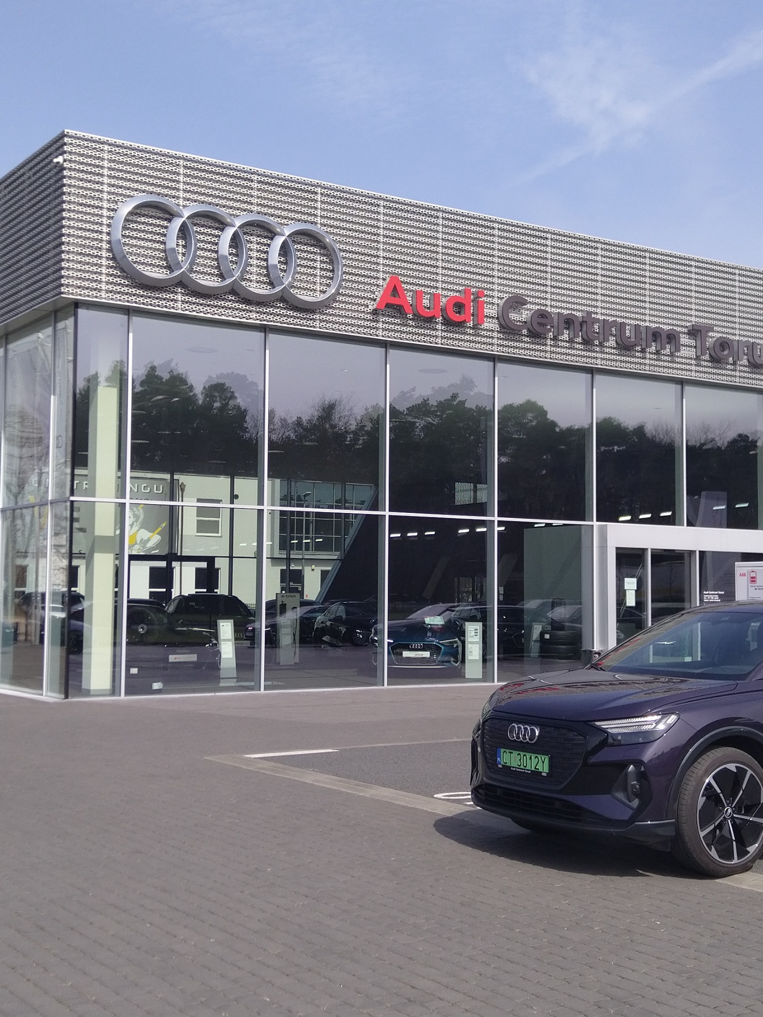 Salon samochodowy Audi Centrum Toruń z zaparkowanym, ciemnofioletowym modelem e-tron na pierwszym planie; widoczne duże logo Audi na fasadzie budynku.