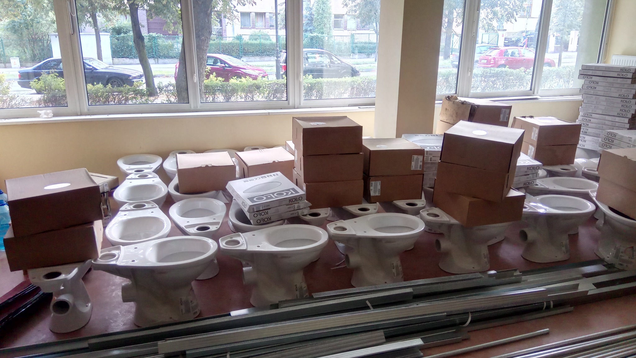 Nowe, białe kompakty WC w magazynie, niektóre jeszcze w kartonach, ułożone w rzędach, widoczne profile metalowe, okno z widokiem na ulicę z samochodami.