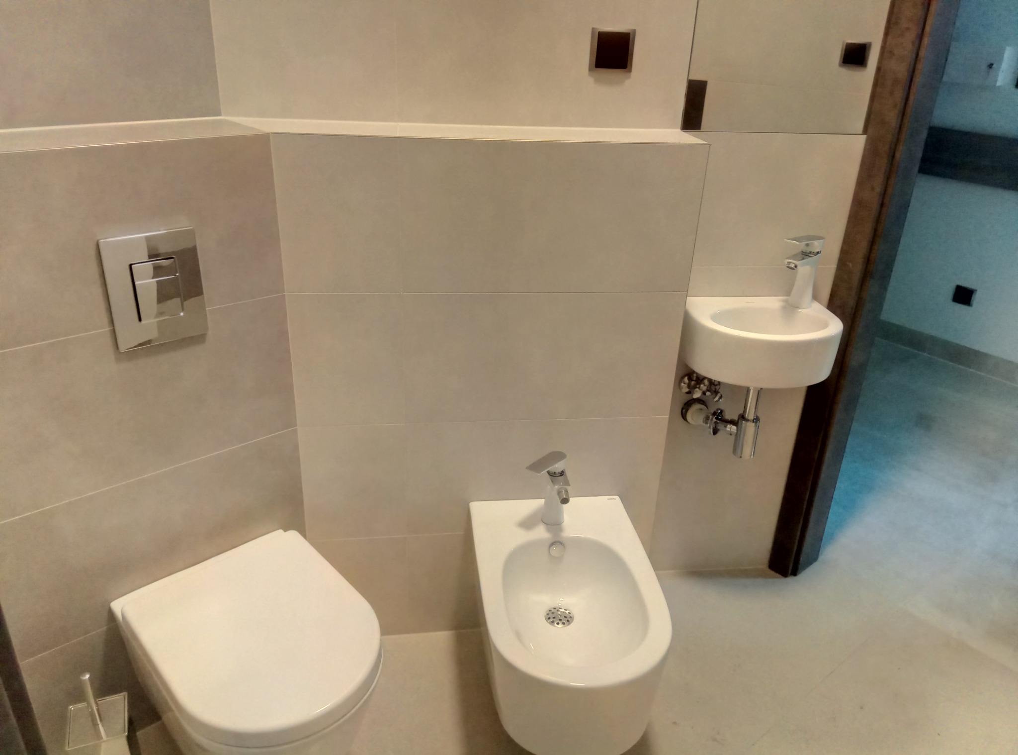 Nowoczesna łazienka z wiszącą toaletą, bidetem i umywalką, wykończona w jasnych, beżowych płytkach ceramicznych z minimalistycznymi akcentami.