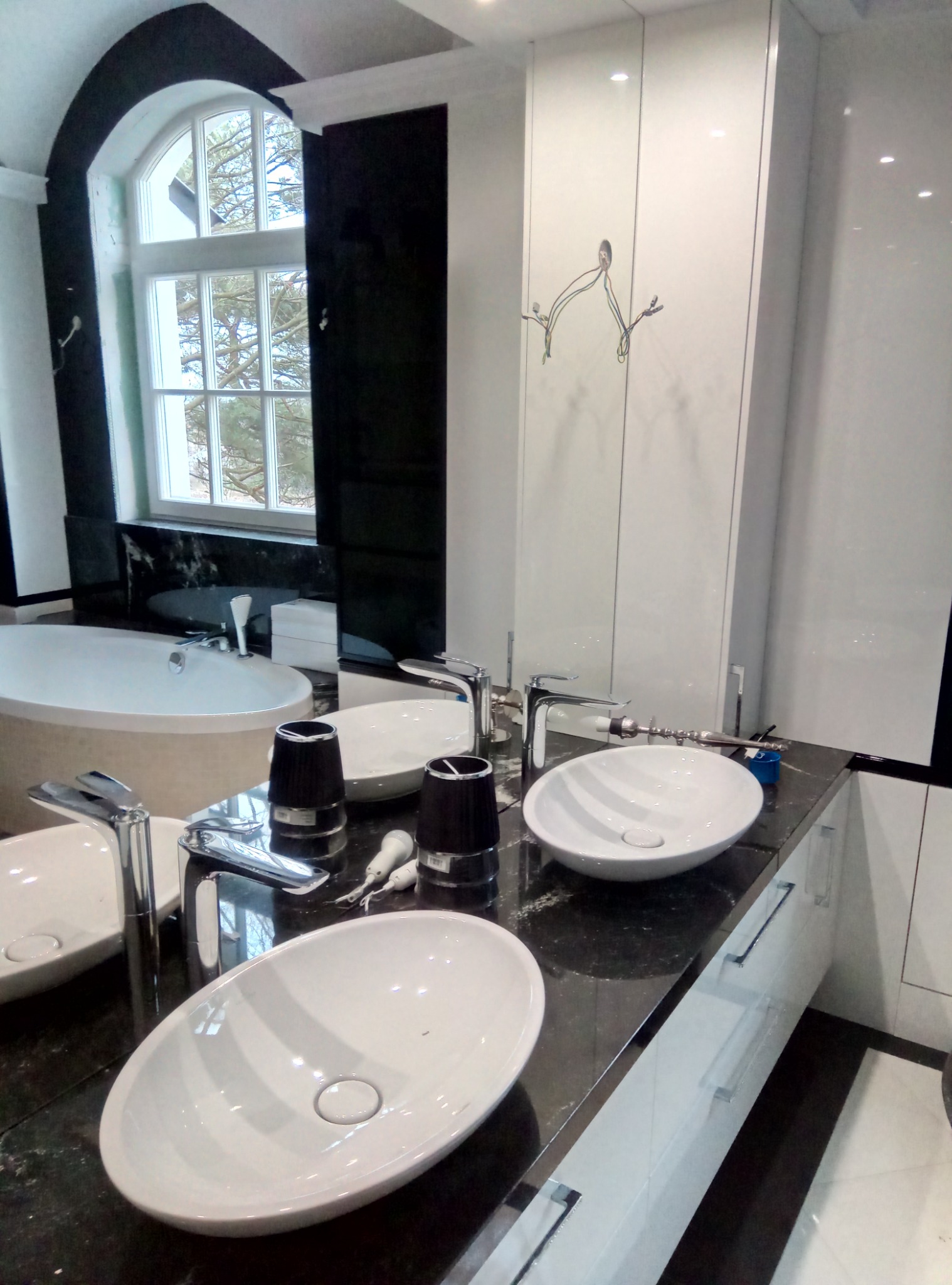 Nowoczesna łazienka z dwiema owalnymi umywalkami nablatowymi, chromowanymi bateriami, czarnym blatem z połyskiem i białymi szafkami. W tle owalne okno z widokiem na drzewa i wanna.