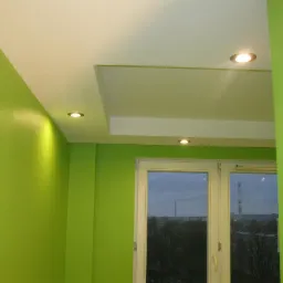 Jasnozielony pokój po remoncie: pomalowane ściany, biały sufit podwieszany z wbudowanymi punktowymi lampami, widok z okna na komin fabryczny.