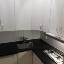 Niewielka kuchnia z białymi szafkami wiszącymi, czarnym blatem, metalowym zlewozmywakiem i dwupalnikową płytą gazową; widoczne białe płytki na ścianie.