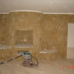 Ściana wykończona płytami z piaskowca w salonie z kominkiem, widoczne półki, odkurzacz i fragment parkietu na podłodze.