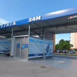 Zewnętrzny widok samoobsługowej myjni samochodowej z napisem 'MYJNIA 24H' na niebieskim dachu, logo Eurowash w prawym górnym rogu, z widocznymi instrukcjami obsługi na ścianach stanowisk...