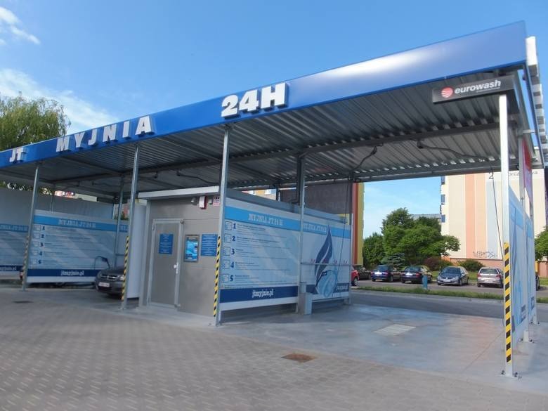 Zewnętrzny widok samoobsługowej myjni samochodowej z napisem 'MYJNIA 24H' na niebieskim dachu, logo Eurowash w prawym górnym rogu, z widocznymi instrukcjami obsługi na ścianach stanowisk...
