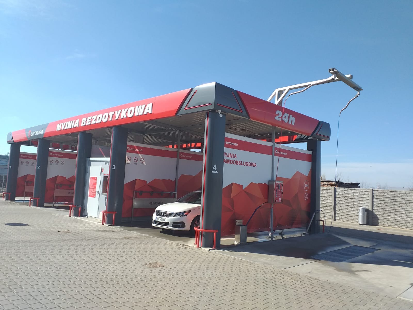 Nowoczesna, bezdotykowa myjnia samochodowa z czterema stanowiskami, oznaczonymi numerami 1-4. Biały samochód osobowy stoi na stanowisku numer 4. Konstrukcja myjni w kolorach czerwieni, bieli...
