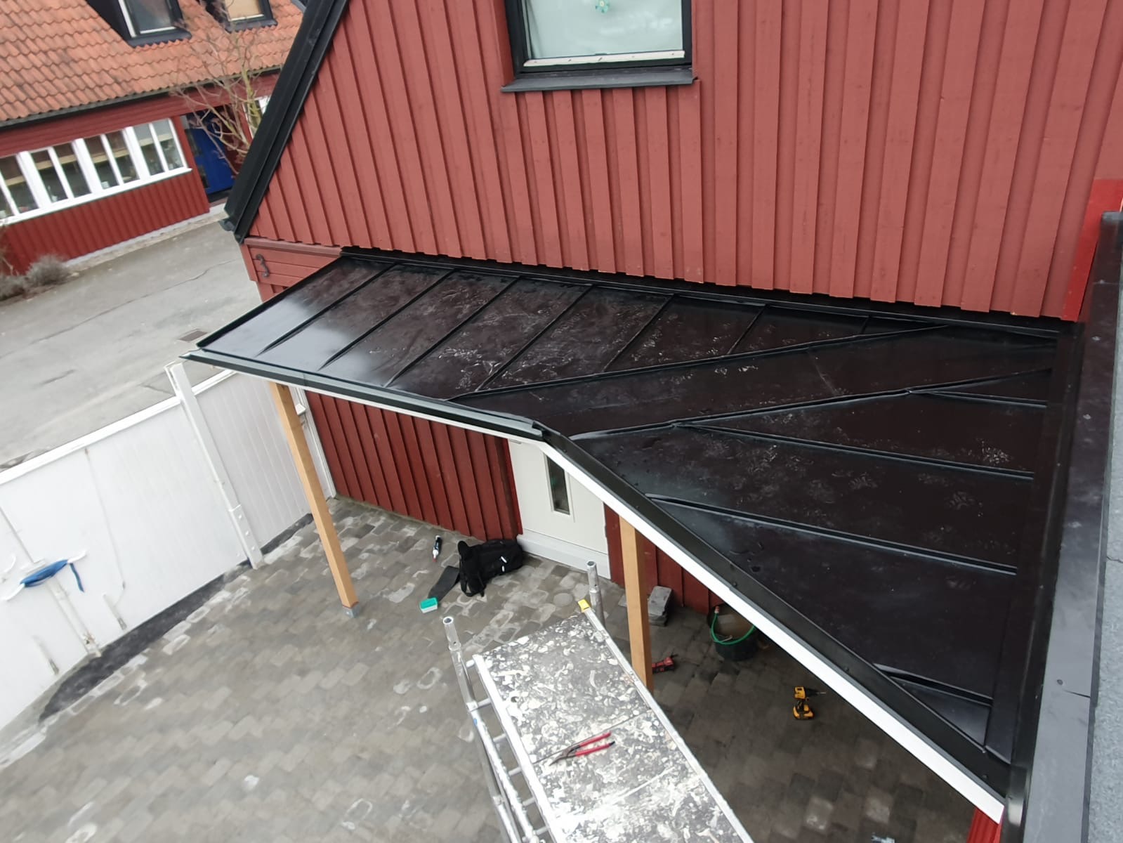 Nowoczesna, czarna konstrukcja zadaszenia nad wejściem do domu z czerwonymi, drewnianymi ścianami, widziana z góry, z widoczną drabiną i narzędziami sugerującymi prace wykończeniowe.