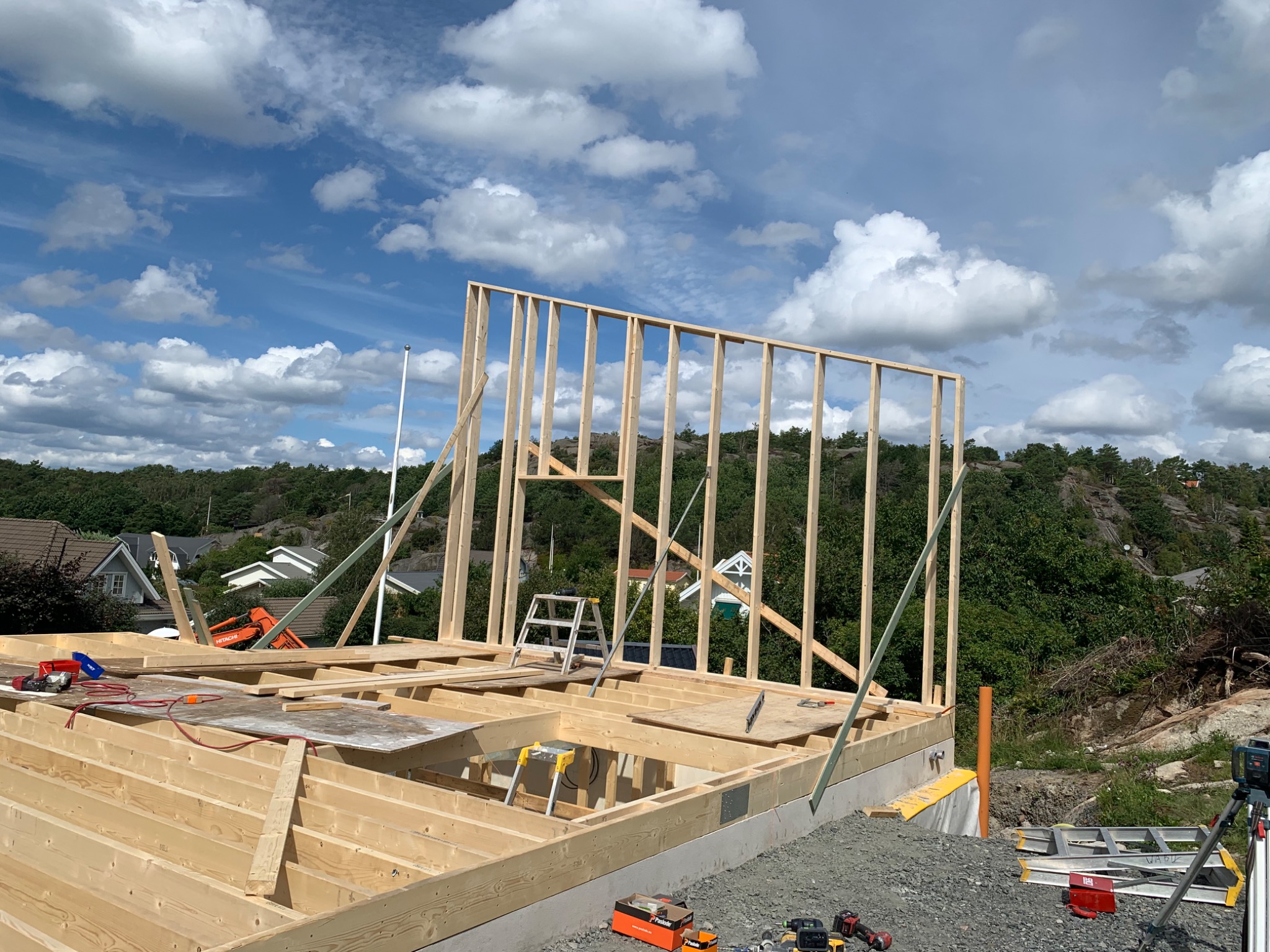 Wznoszenie drewnianej konstrukcji szkieletowej domu na fundamencie, widoczne narzędzia, poziomica laserowa i drabina, w tle zabudowa jednorodzinna i zalesione wzgórze pod błękitnym niebem z obłokami.