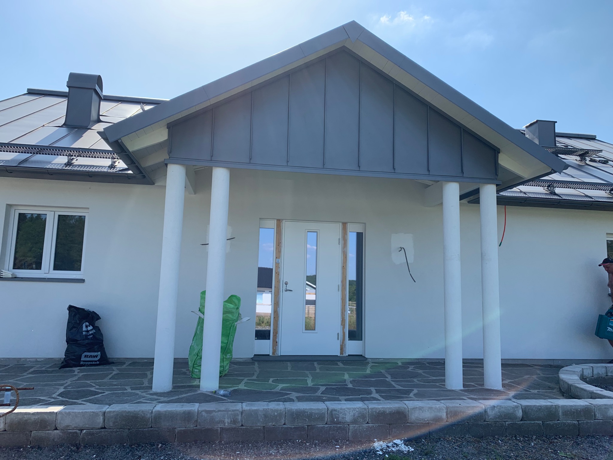 Nowoczesny dom z szarą blachą na dachu i nad wejściem, białe kolumny podtrzymujące zadaszenie wejścia, widoczne worki z odpadami budowlanymi.