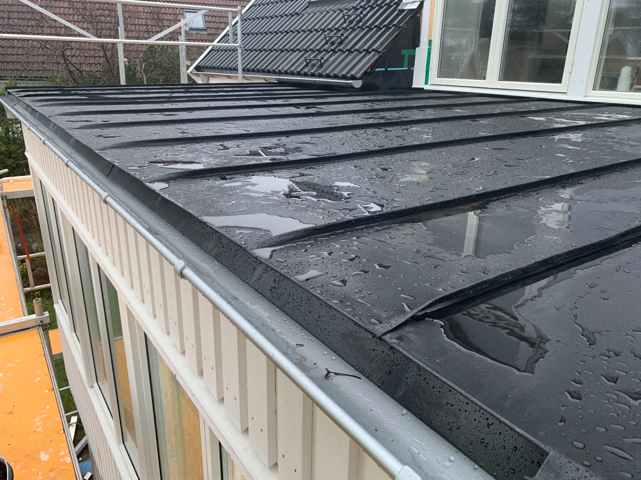 Metalowy dach po deszczu, widoczna rynna, krople wody na powierzchni, biała elewacja budynku z oknami.