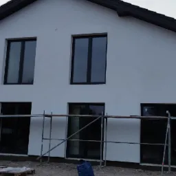 Nowoczesny dom z białą elewacją i czarnymi oknami, w trakcie prac wykończeniowych, widoczne rusztowania i materiały budowlane.