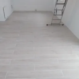 Ułożone jasne, podłużne płytki podłogowe imitujące drewno w pustym pomieszczeniu z białą ścianą i grzejnikiem, drabina oparta o ścianę, okno w tle.