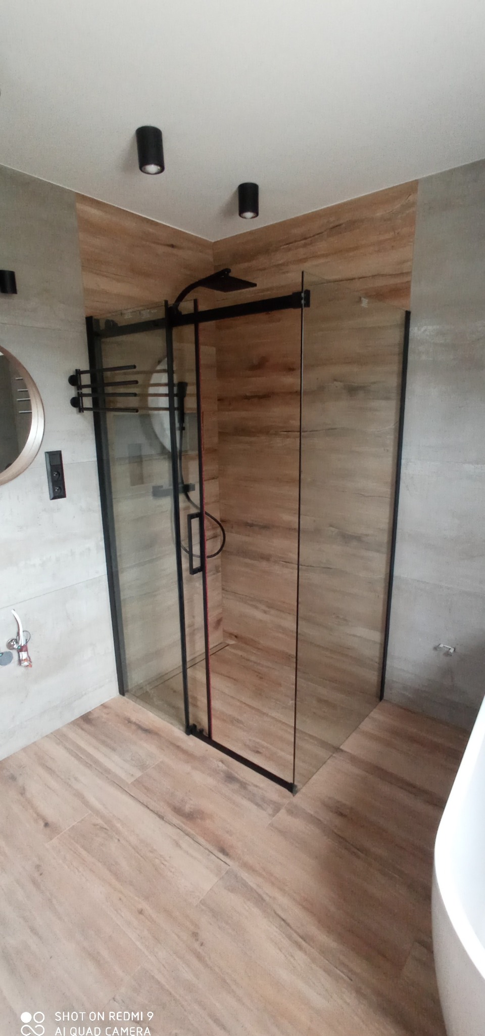 Nowoczesna kabina prysznicowa z czarnymi profilami i deszczownicą, wykończona płytkami imitującymi drewno, z widocznym okrągłym lustrem i punktowym oświetleniem sufitowym.