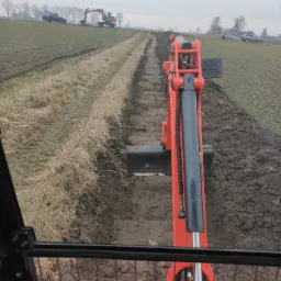 Widok z kabiny pomarańczowej minikoparki podczas kopania rowu w ziemi, w tle widoczna inna koparka i samochody na polu, pochmurne niebo.