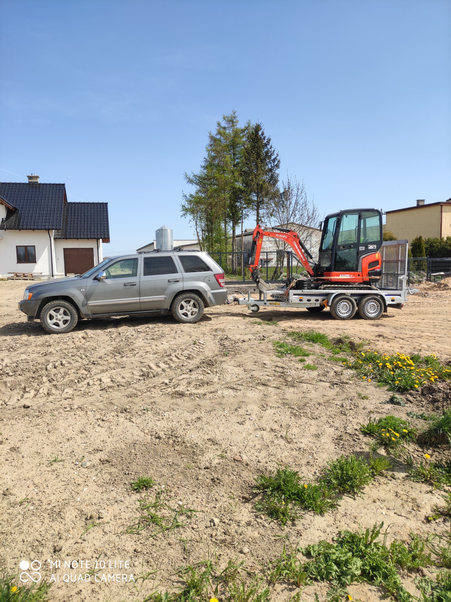 Szary samochód terenowy ciągnie na przyczepie pomarańczową mini koparkę Kubota na plac budowy z widocznymi śladami opon na ubitej ziemi, w tle dom jednorodzinny i drzewa.