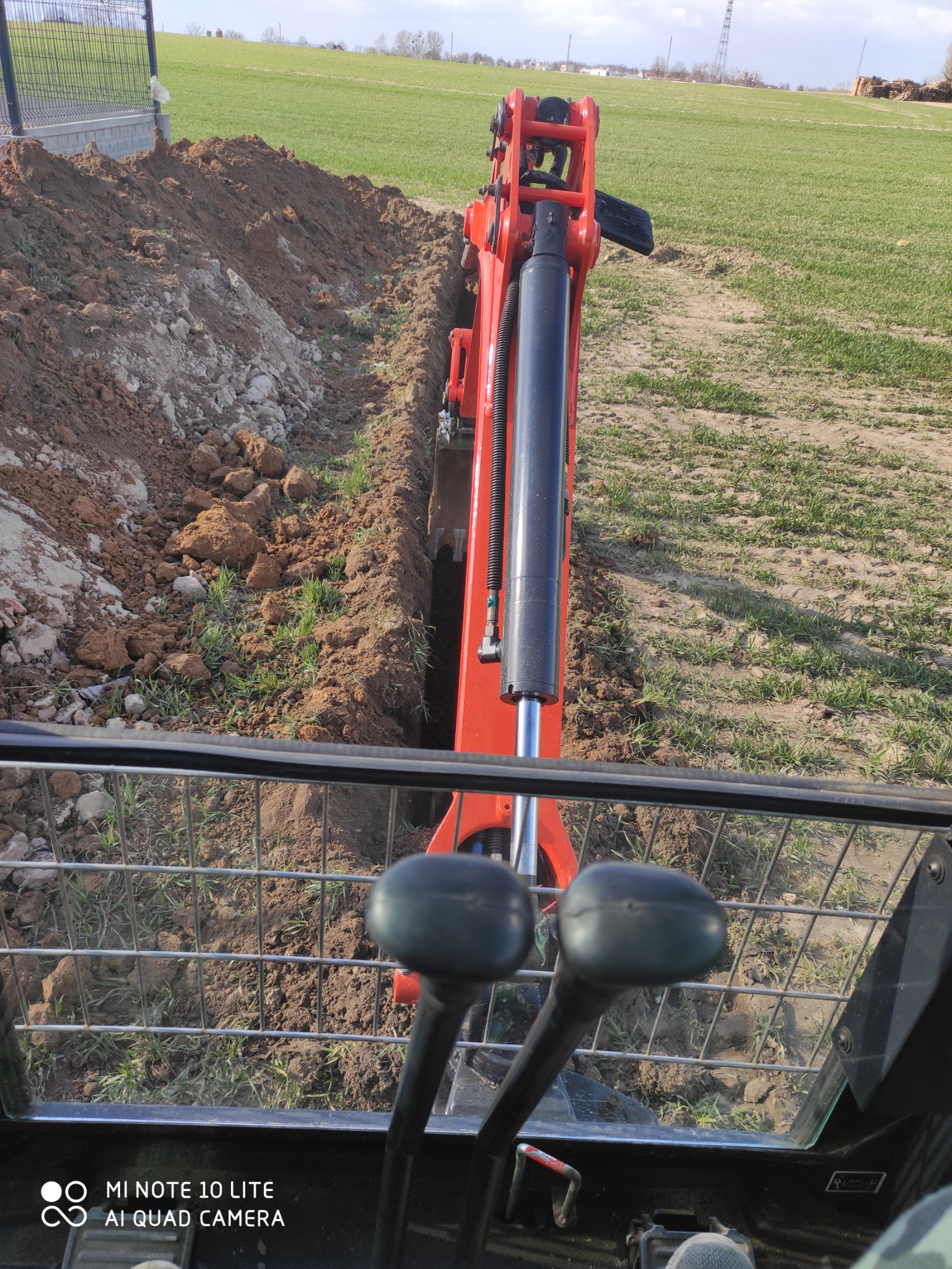 Widok z kabiny operatora na czerwoną mini-koparkę podczas wykonywania wąskiego wykopu w ziemi na polu. Ziemia usypana po obu stronach wykopu. W tle zielone pole i słupy energetyczne.