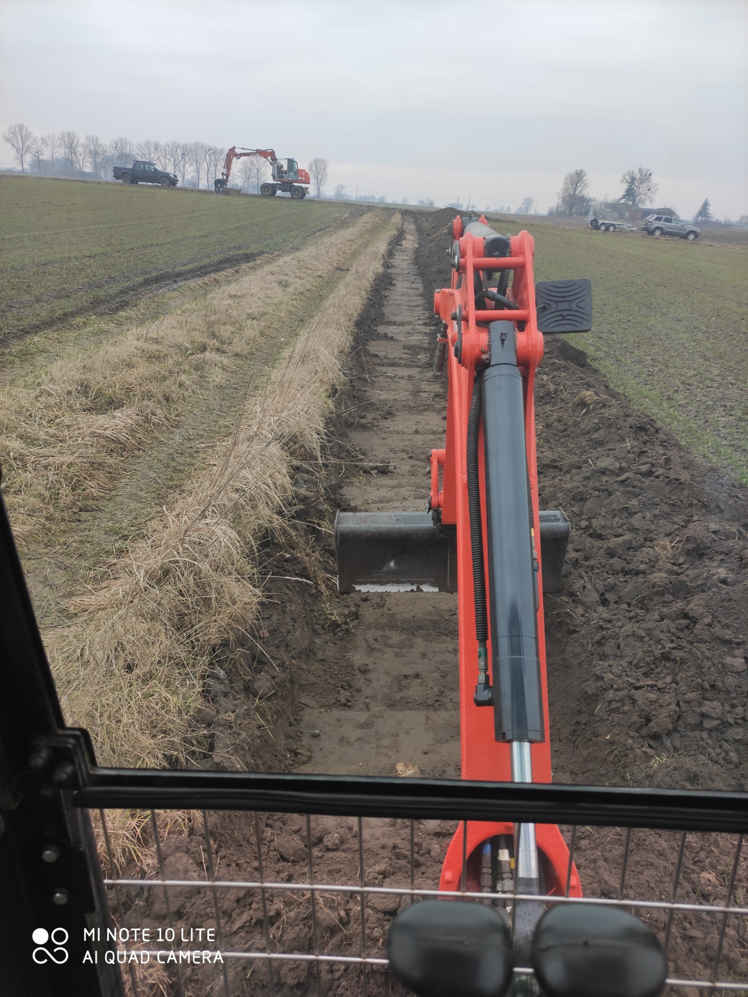 Widok z kabiny pomarańczowej minikoparki podczas kopania rowu w ziemi, w tle widoczna inna koparka i samochody na polu, pochmurne niebo.