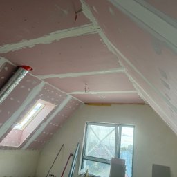 Art-Gips Patryk Rentkowski - Poddasze z oknem dachowym w trakcie wykańczania: różowe płyty karton-gips na skosach, szpachlowane łączenia. Widoczne narzędzia i fragment okna.
