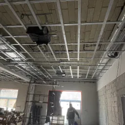 Montaż stelaża sufitowego w Rymań. Widoczne profile metalowe, izolacja i instalacje. W tle okno i pracownik w bluzie. Surowy stan wykończenia wnętrza.