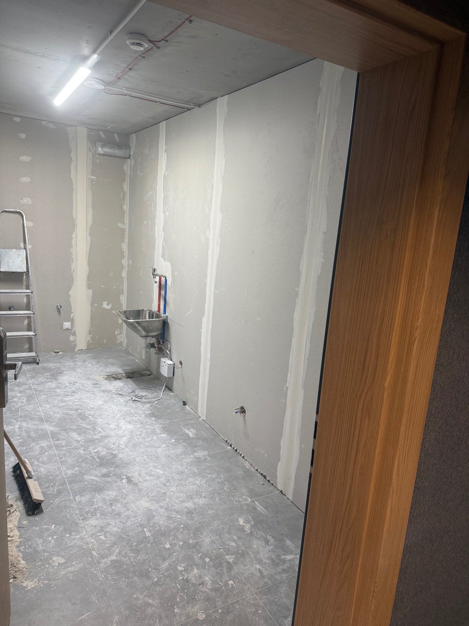 Wnętrze w trakcie prac wykończeniowych: ściany z płyt karton-gips, instalacja umywalki, drabina i szczotka. Widok zza drewnianej ościeżnicy.