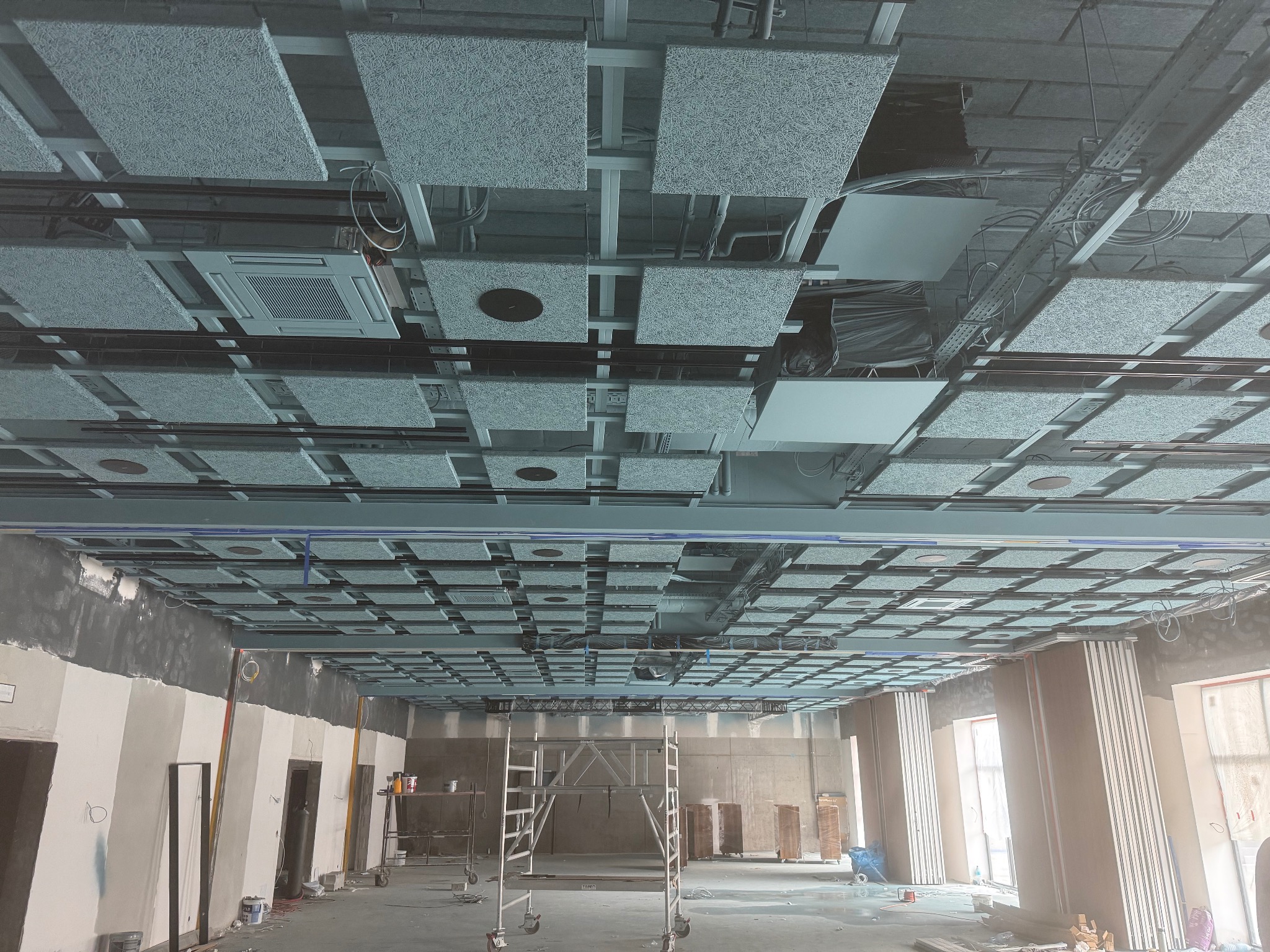 Montaż sufitu akustycznego z kwadratowych paneli w pomieszczeniu z widoczną konstrukcją nośną i instalacjami. Widoczne rusztowanie i przygotowane ściany.