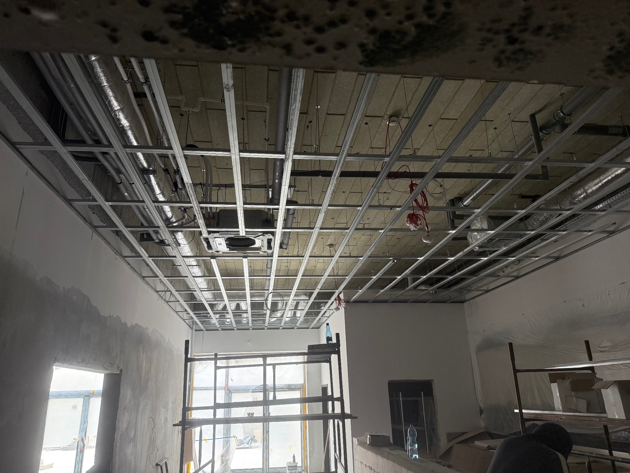 Montaż metalowej konstrukcji sufitu podwieszanego z widocznymi instalacjami, w surowym wnętrzu z oknami i rusztowaniami. Etap prac wykończeniowych.